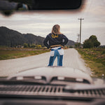 Person standing on a road with mountains in the background, viewed from inside a vehicle wearing Soul Sister since 1969.