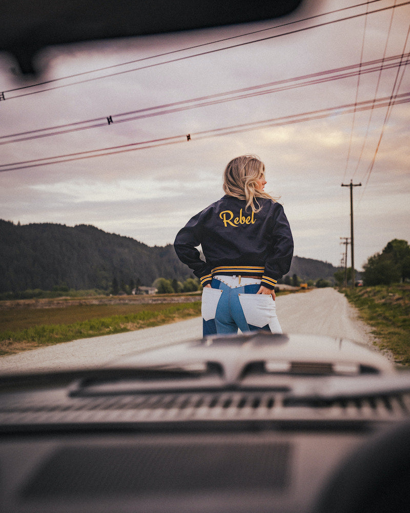 Person wearing a jacket with 'Rebel' on it standing on a road with mountains in the background wearing Soul Sister since 1969