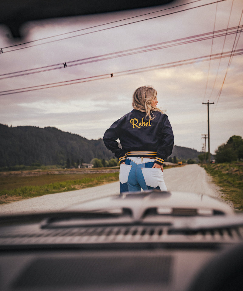 Person wearing a jacket with 'Rebel' on it standing on a road with mountains in the background wearing Soul Sister since 1969