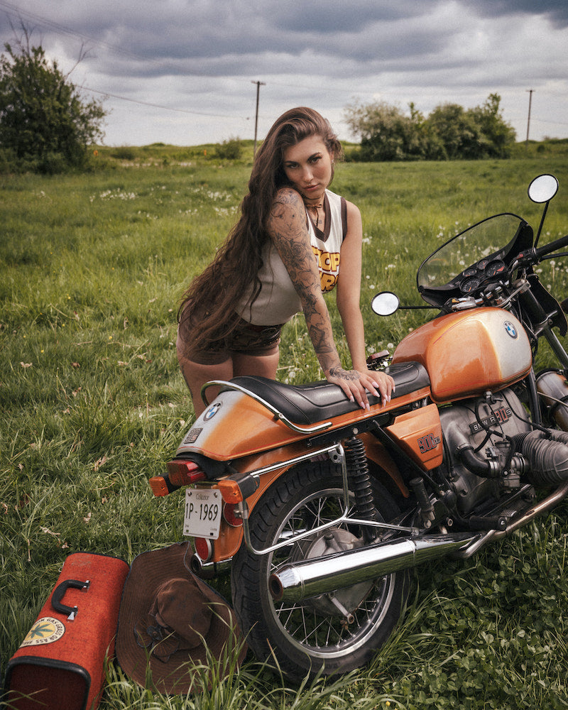 Woman with tattoos sitting on an orange motorcycle in a grassy field wearing clothing designed by Soul Sister since 1969
