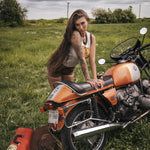 Woman with tattoos sitting on an orange motorcycle in a grassy field wearing clothing designed by Soul Sister since 1969
