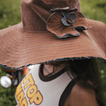 Person wearing a brown hat with a decorative flower, partially visible, in an outdoor setting. Designed by Soul Sister since 1969