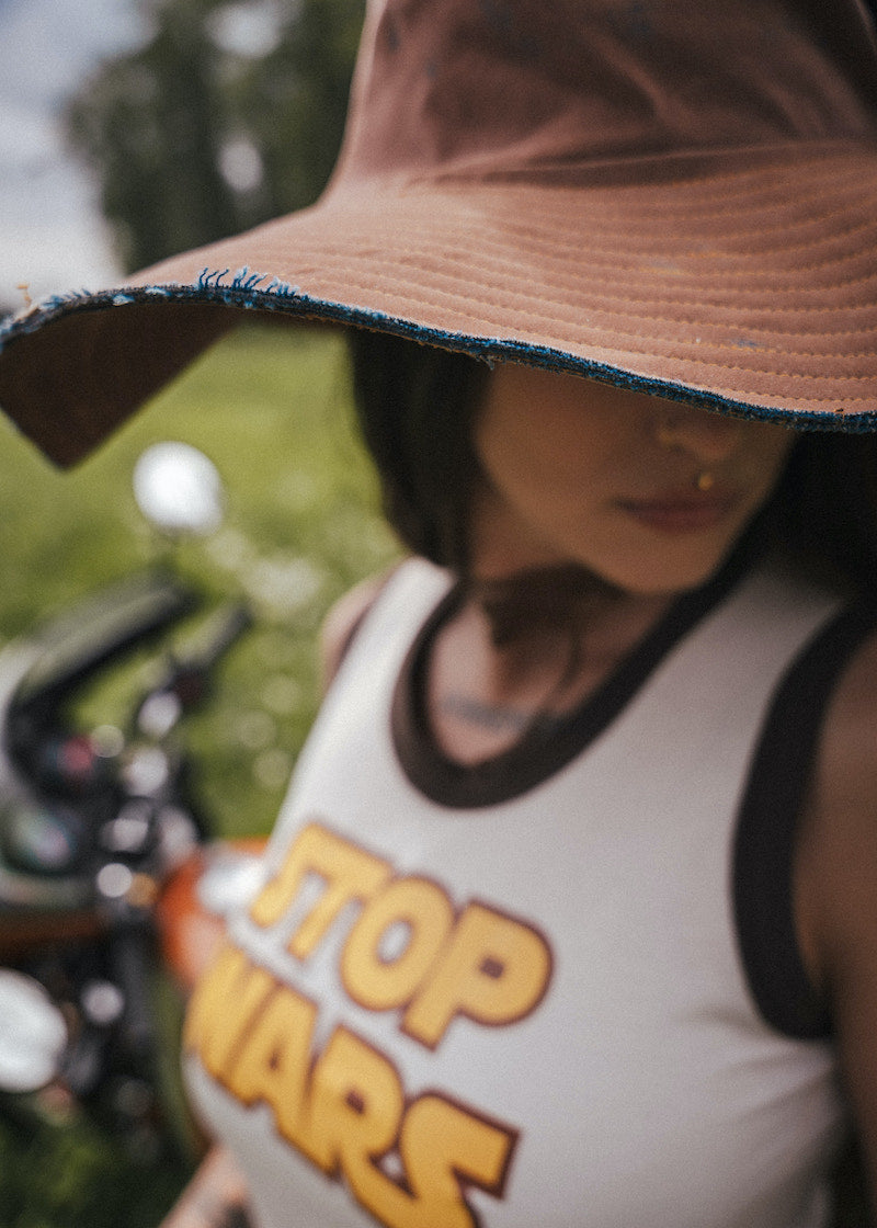 Person wearing a tank top with 'STOP WARS' text, standing outdoors wearing an oversized hat 