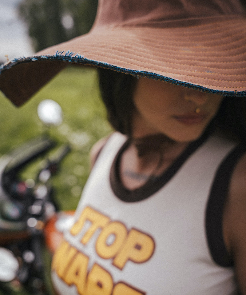 Person wearing a tank top with 'STOP WARS' text, standing outdoors wearing an oversized hat 