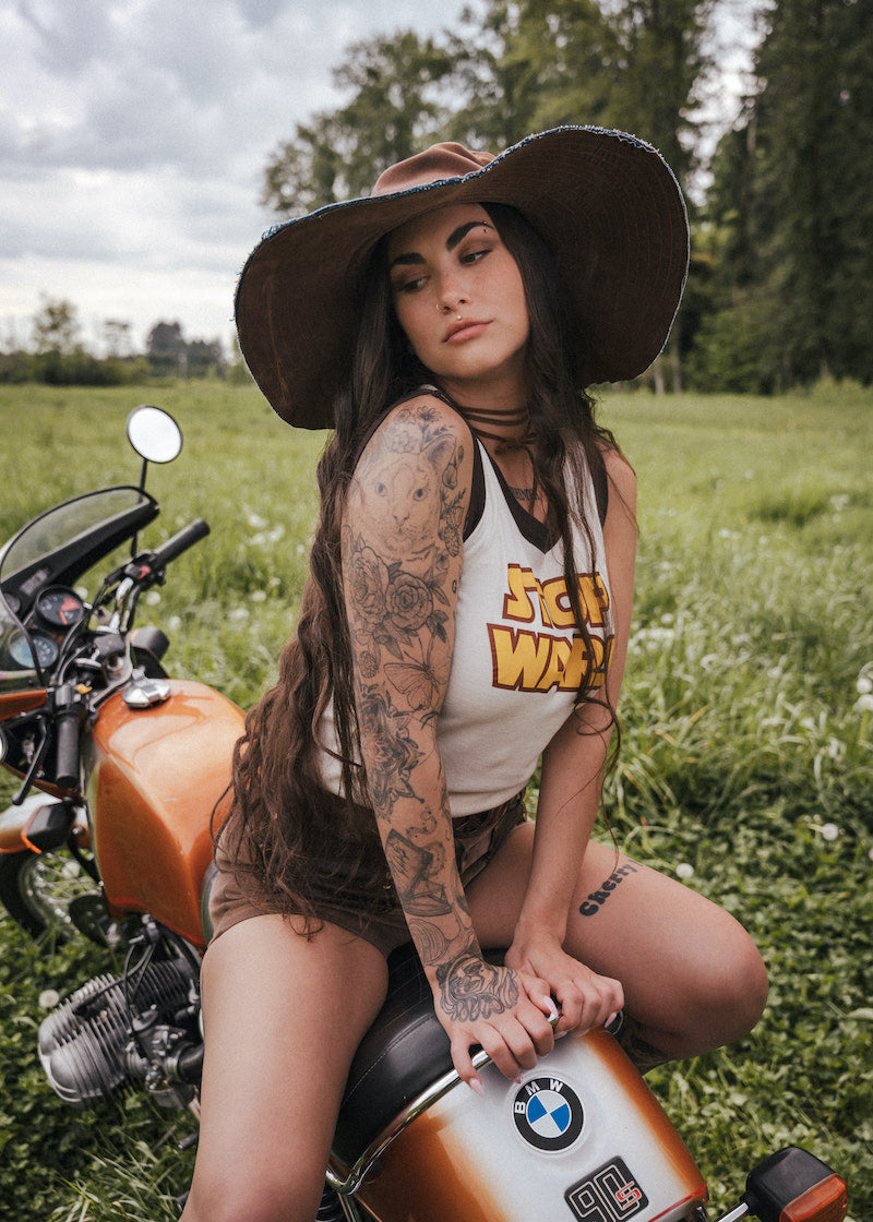 Woman with tattoos sitting on a motorcycle in a field wearing a hat and clothing Designed by Soul Sister since 1969