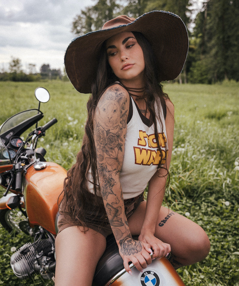 Woman with tattoos sitting on a motorcycle in a field wearing a hat and clothing Designed by Soul Sister since 1969