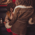 Person wearing a brown coat with fur trim interacting with another person in a dimly lit room.