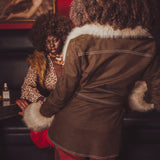 Person wearing a brown coat with fur trim interacting with another person in a dimly lit room.