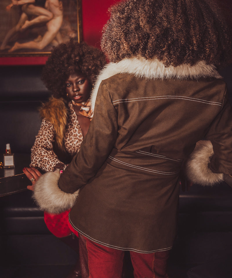 Person wearing a brown coat with fur trim interacting with another person in a dimly lit room.