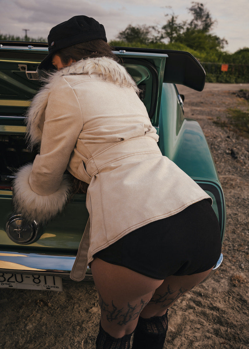 Person in a beige coat and black hat leaning into a vintage car with a fur-lined hood wears clothing by Soul Sister since 1969.