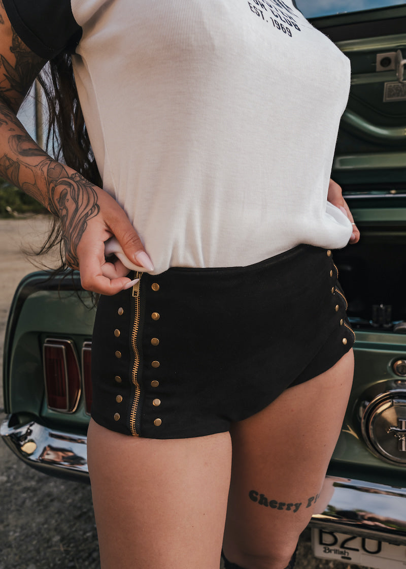 Person wearing a white top and black shorts with gold buttons, standing next to a vintage car wears clothing by Soul Sister since 1969.