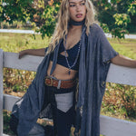 A woman wearing a knit crochet lace robe in gray, with a skinny scarf, paired with jeans and standing by a fence in a natural outdoor setting.