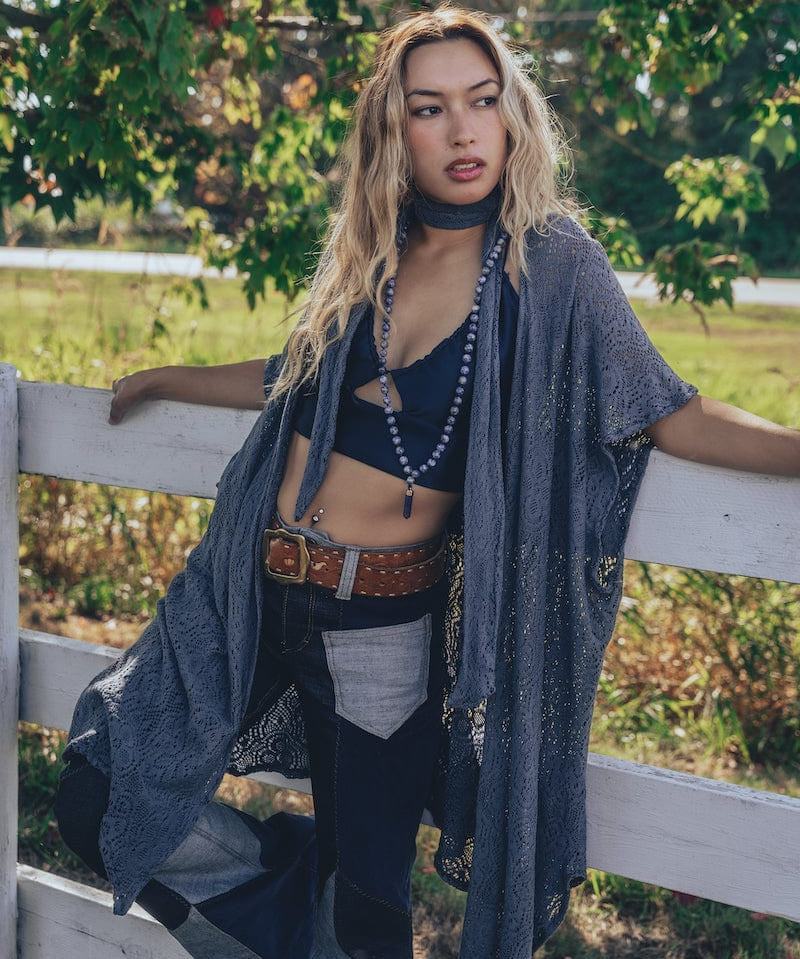 A woman wearing a knit crochet lace robe in gray, with a skinny scarf, paired with jeans and standing by a fence in a natural outdoor setting.