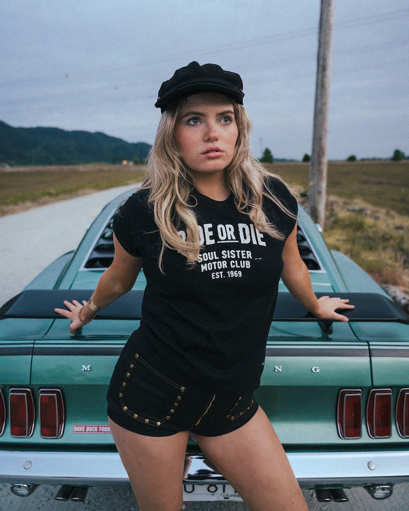 Woman wearing a black 'De or Die' t-shirt standing next to a vintage car on a road Ride or Die Motor Club wearing soul sister since 1969.
