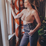 Woman standing by a window with patterned curtains, wearing a beige top and blue jeans.