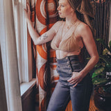 Woman standing by a window with patterned curtains, wearing a beige top and blue jeans.