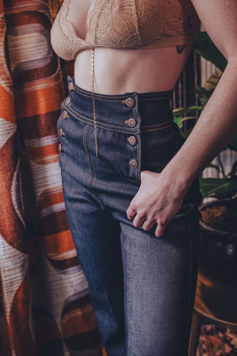 Person wearing a beige bralette and high-waisted blue jeans against a patterned curtain background.