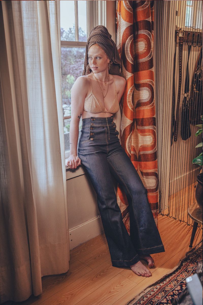 Woman in a beige top and dark pants standing by a window with colorful curtains.