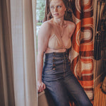 Woman in a beige top and blue jeans standing by a window with patterned curtains.