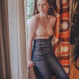 Woman in a beige top and blue jeans standing by a window with patterned curtains.