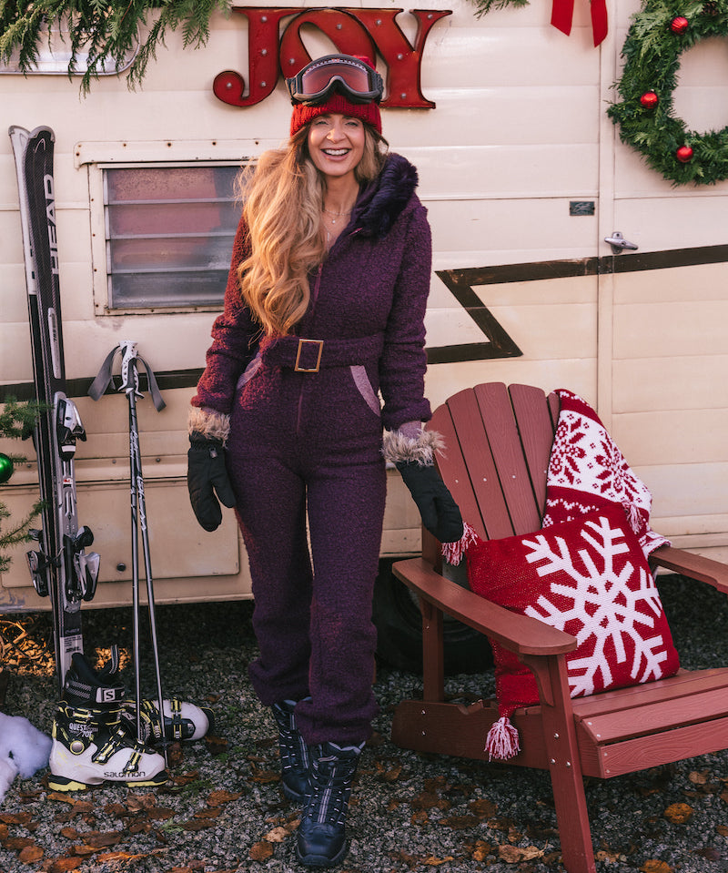 Woman in a purple snowsuit standing next to a wooden chair with a red and white blanket, in front of a trailer with Christmas decorations.