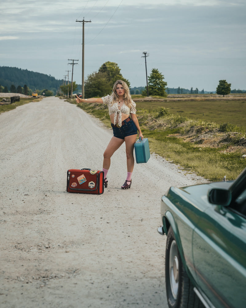 Woman standing on a dirt road with suitcases, hitchhiking wearing Soul Sister since 1969..