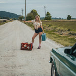 Woman standing on a dirt road with suitcases, hitchhiking wearing Soul Sister since 1969..