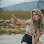 Woman sitting on a gravel road with mountains in the background, giving a thumbs-up wearing Soul Sister since 1969.