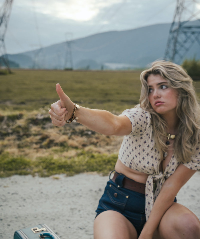 Woman sitting on a gravel road with mountains in the background, giving a thumbs-up wearing Soul Sister since 1969.