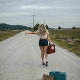 Woman standing on a road with suitcases and a sign, indicating a journey or travel theme wearing Soul Sister since 1969..