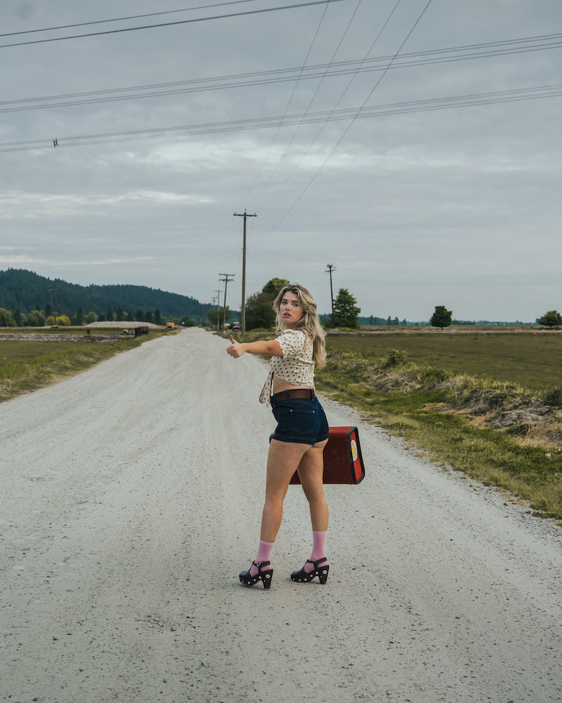 Woman hitchhiking on a rural road with a suitcase wearing Soul Sister since 1969.
