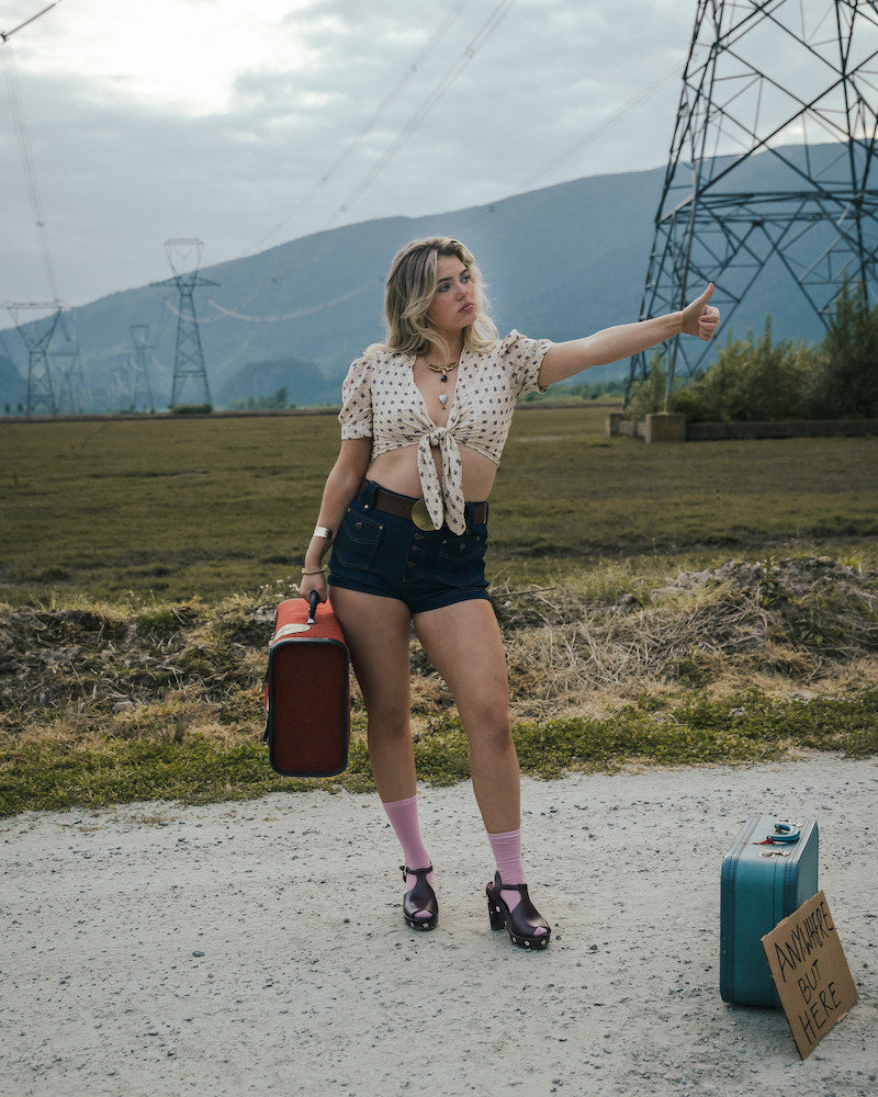 Woman hitchhiking on a road with suitcases and a sign in an open field wearing Soul Sister since 1969.
