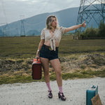 Woman hitchhiking on a road with suitcases and a sign in an open field wearing Soul Sister since 1969.