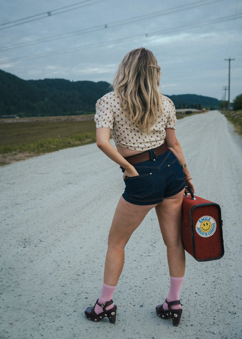 Woman standing on a road holding a red suitcase with a smiley face, wearing a polka dot shirt and denim shorts. wearing Soul Sister since 1969.