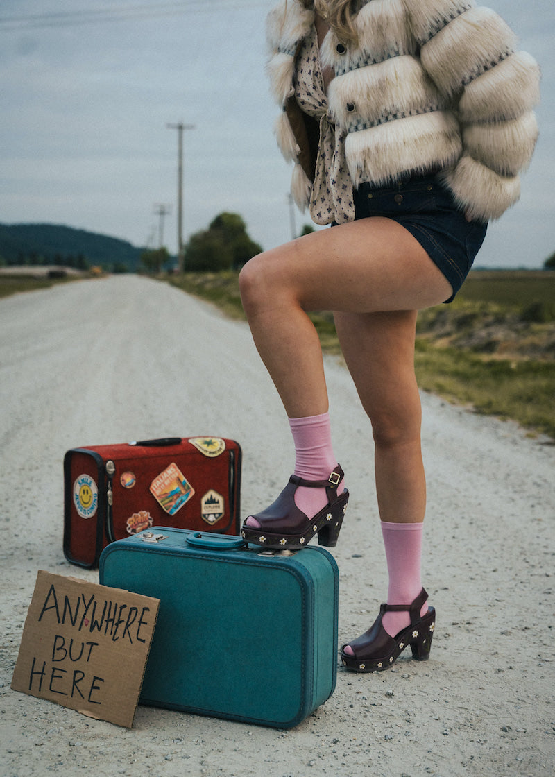 Person standing on a road with suitcases and a sign reading 'Anywhere But Here' wearing a fluffy coat and pink socks wearing Soul Sister since 1969..