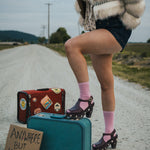 Person standing on a road with suitcases and a sign reading 'Anywhere But Here' wearing a fluffy coat and pink socks wearing soul sister since 1969.