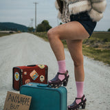 Person standing on a road with suitcases and a sign reading 'Anywhere But Here' wearing a fluffy coat and pink socks wearing soul sister since 1969.