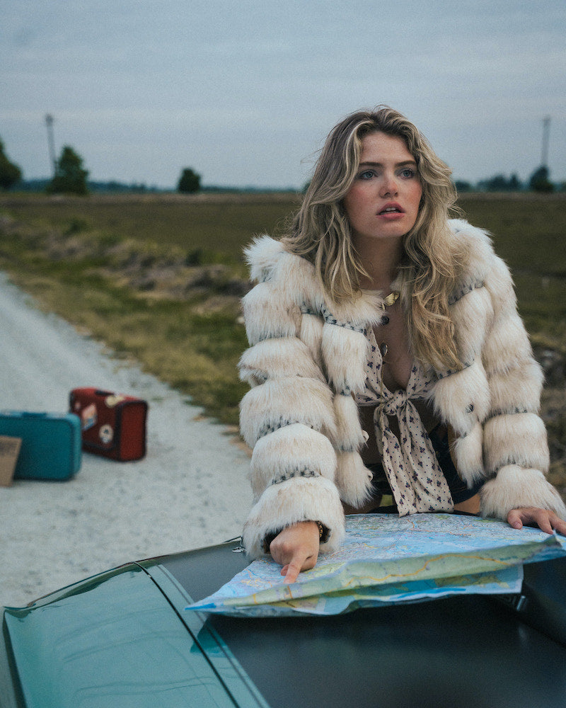 Woman in a fur coat sitting on a car hood with a map, surrounded by suitcases in an open field wearing soul sister since 1969.