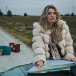 Woman in a fur coat sitting on a car hood with a map, surrounded by suitcases in an open field wearing soul sister since 1969.