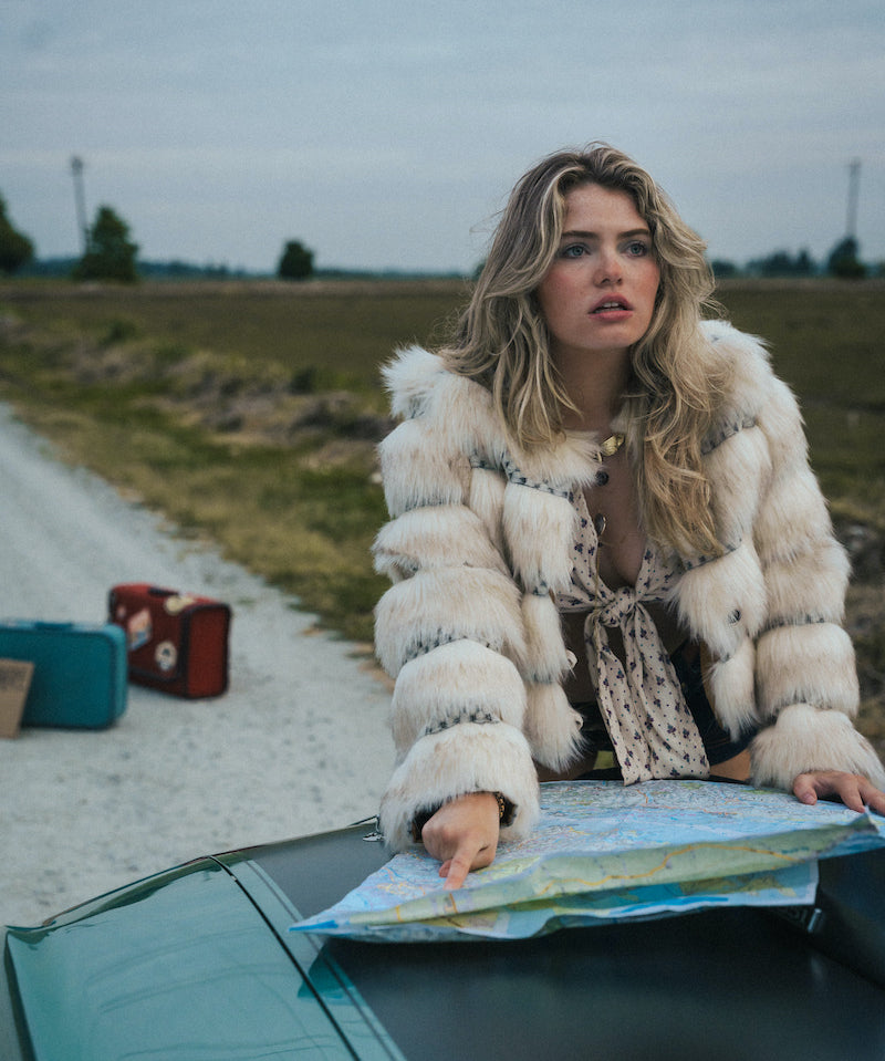 Woman in a fur coat sitting on a car hood with a map, surrounded by suitcases in an open field wearing soul sister since 1969.