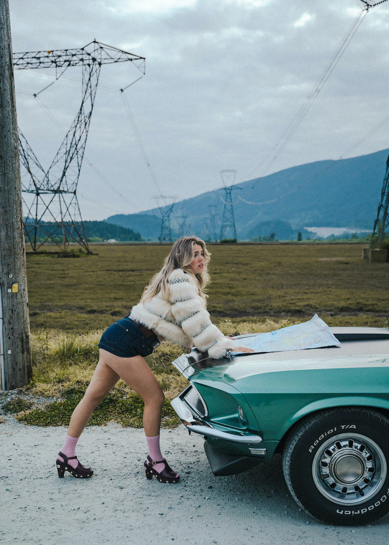 Woman in a puffer vest leaning on a vintage car in an open field with power lines and mountains in the background. wearing soul sister since 1969