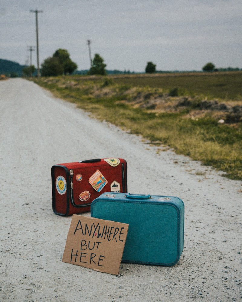 Two suitcases on a rural road with a sign reading 'Anywhere But Here'.