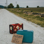 Two suitcases on a rural road with a sign reading 'Anywhere But Here'.