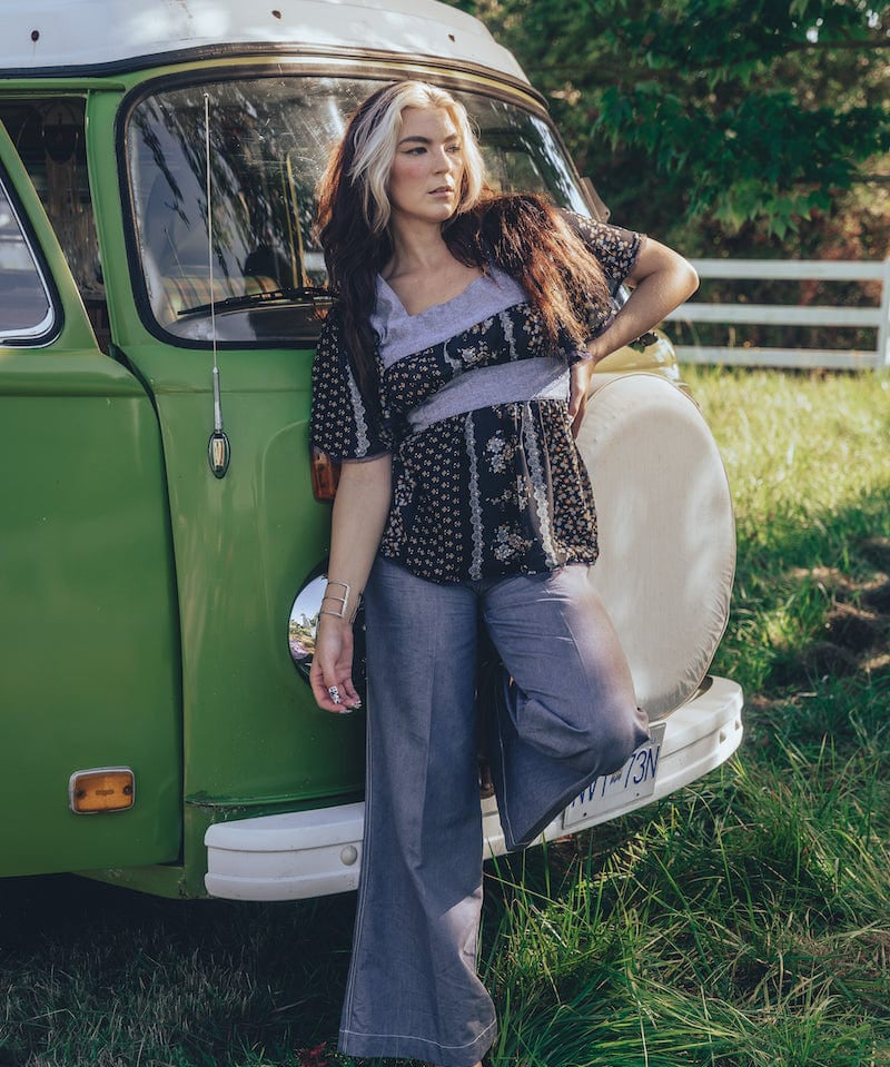 A woman standing beside a green van, wearing a black and white floral print tunic top with long sleeves, paired with gray pants.