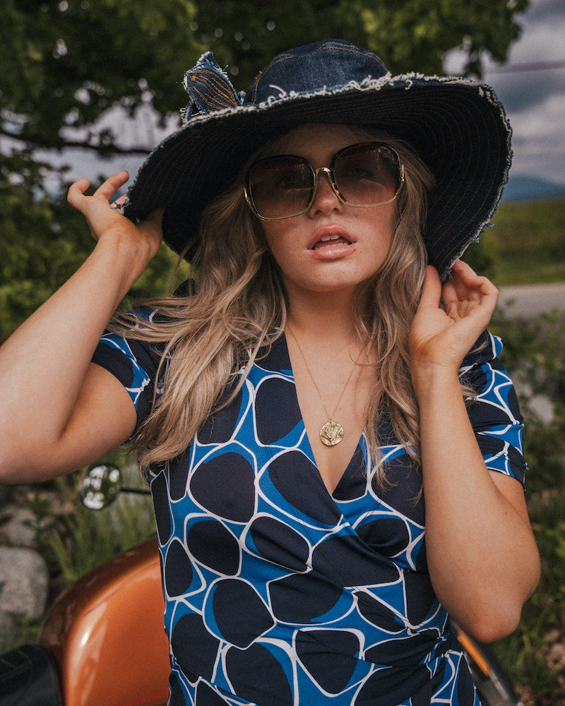 Woman wearing a blue and black patterned dress, large sun hat, and sunglasses outdoors wearing Soul Sister since 1969..
