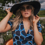 Woman wearing a blue and black patterned dress, large sun hat, and sunglasses outdoors wearing Soul Sister since 1969..