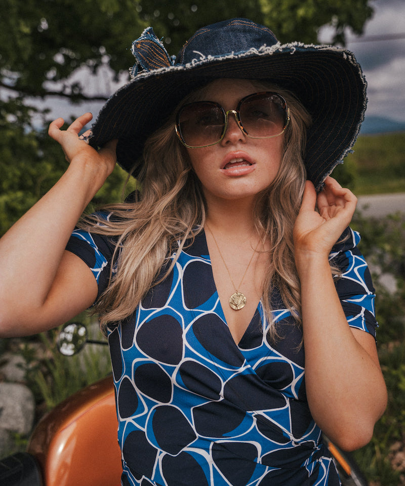 Woman wearing a blue and black patterned dress, large sun hat, and sunglasses outdoors wearing Soul Sister since 1969..