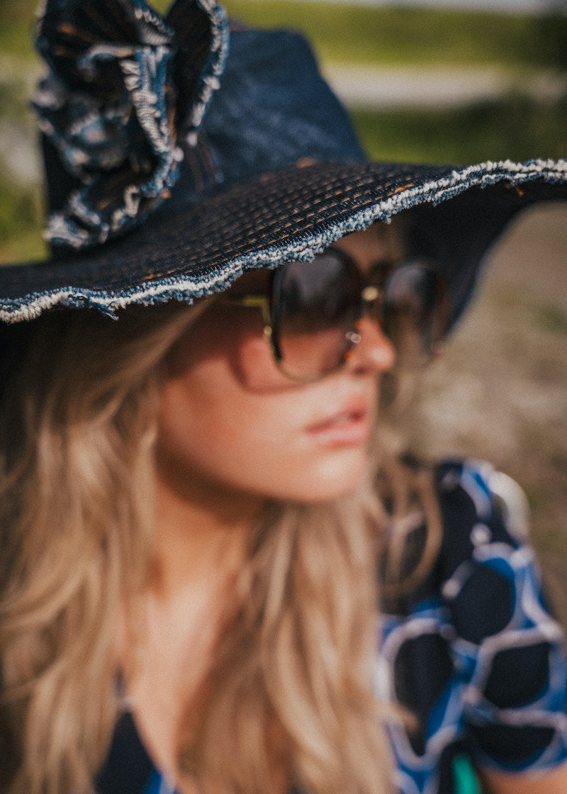 Person wearing a black hat with decorative trim and sunglasses, with a blurred natural background wearing Soul Sister since 1969.