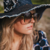 Person wearing a black hat with decorative trim and sunglasses, with a blurred natural background wearing Soul Sister since 1969.