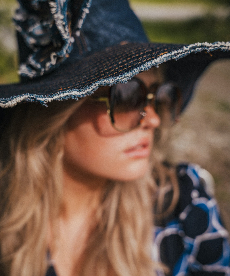 Person wearing a black hat with decorative trim and sunglasses, with a blurred natural background wearing Soul Sister since 1969.